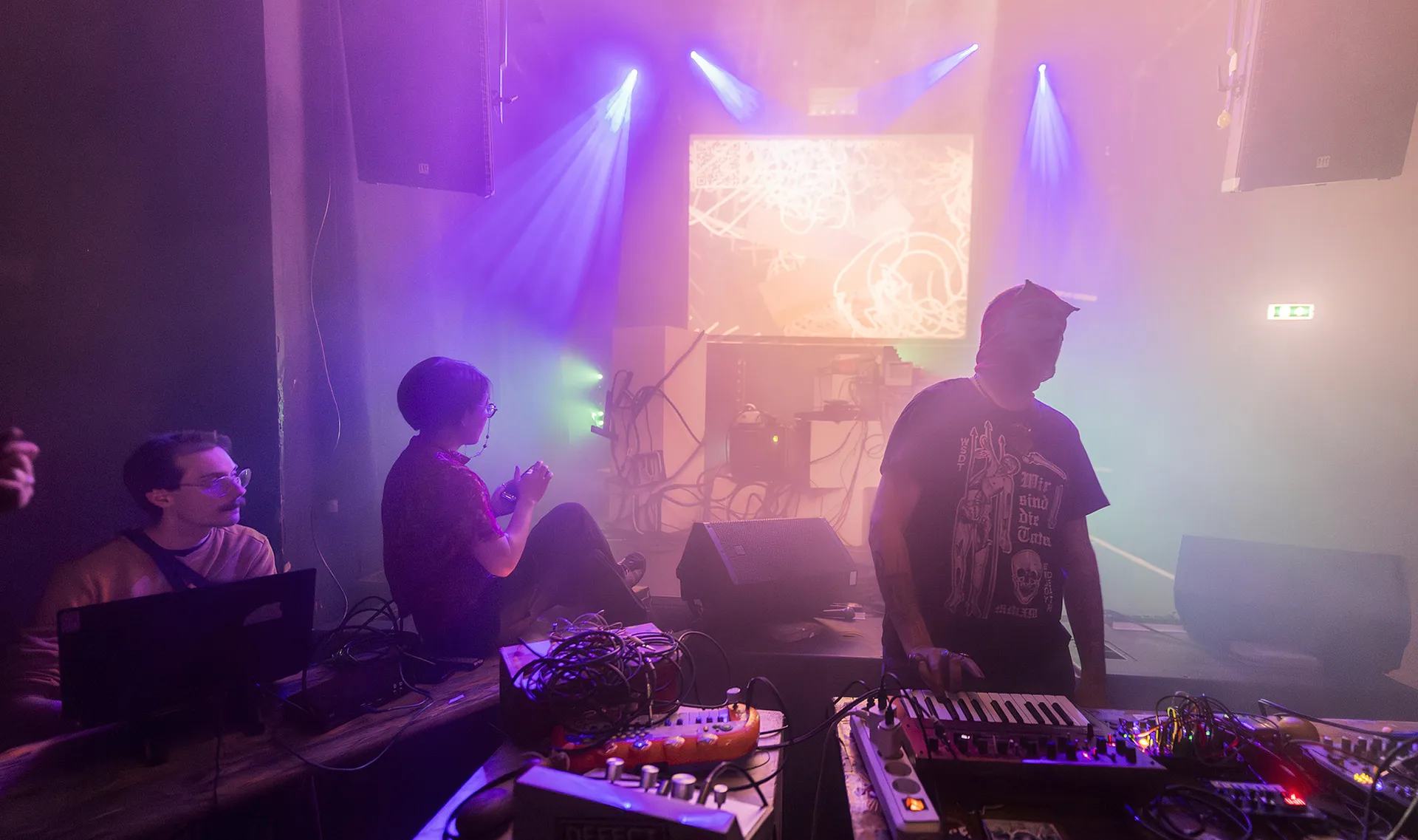 A foggy club stage with some electronics synths and projections in the background.